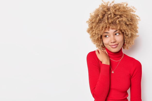 Gratis foto blij krullend haired jonge vrouw houdt hand op schouder draagt comfortabele rode coltrui denkt aan iets aangenaams geïsoleerd op witte achtergrond met kopie ruimte voor uw advertentie