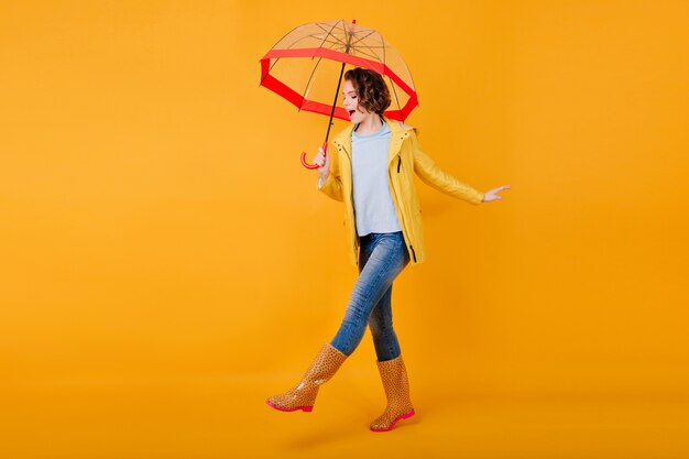 Blij krullend meisje in jeans grappige dansende trendy parasol te houden. Studio portret van geïnspireerde jonge vrouw in rubberen schoenen gek rond op felgele muur en glimlachen.