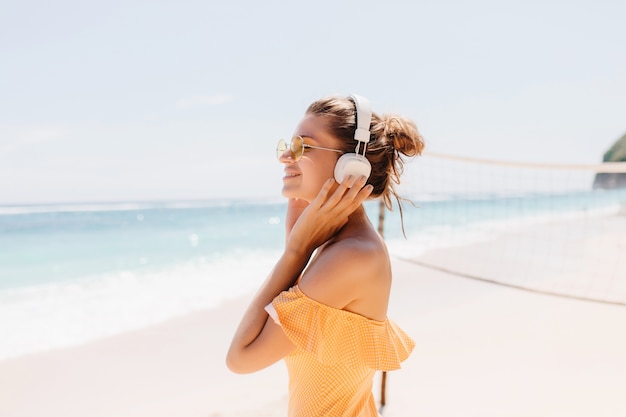 Blij lachende vrouw met gebruinde huid poseren op het strand met charmante glimlach. Outdoor Portret van enthousiast meisje draagt grote witte koptelefoon tijdens het koelen aan de kust van de oceaan.