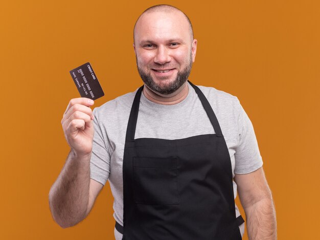 Blij mannelijke kapper van middelbare leeftijd in uniform met creditcard geïsoleerd op oranje muur