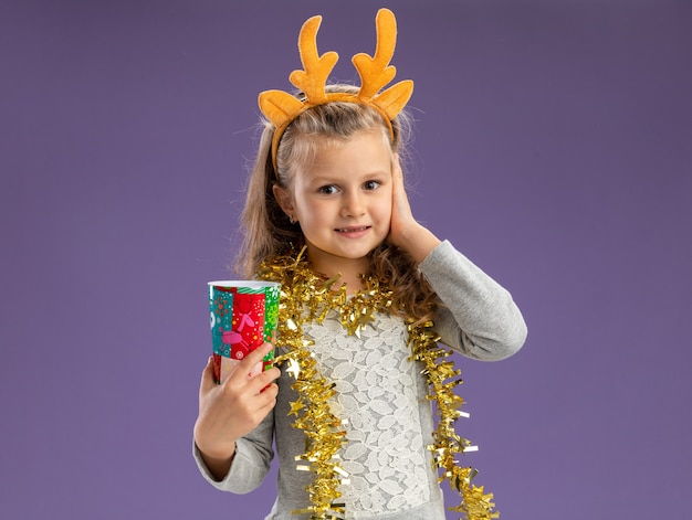 Blij meisje dragen kerst haar hoepel met garland op nek houden kerst beker hand op Wang geïsoleerd op blauwe achtergrond met kopie ruimte