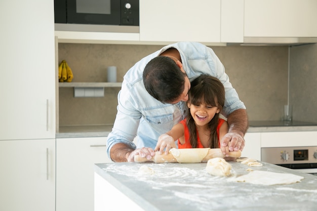 Blij meisje en haar vader met plezier in de keuken tijdens het rollen en kneden van deeg.