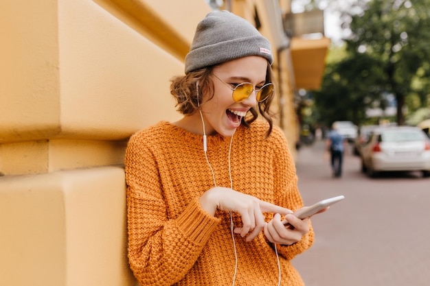 Blij meisje in gezellige outfit met plezier buiten wachtende vriend om samen te lopen. portret van een mooi vrouwelijk model in gele trui die lacht terwijl ze mobiele mail controleert op de achtergrond van de wazige straat.