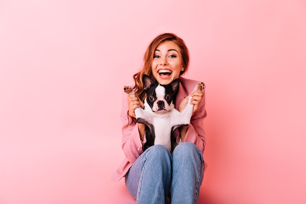 Blij meisje in spijkerbroek spelen met grappige kleine hond. Indoor portret van opgewonden gember dame met krullend kapsel tijd doorbrengen met haar puppy.