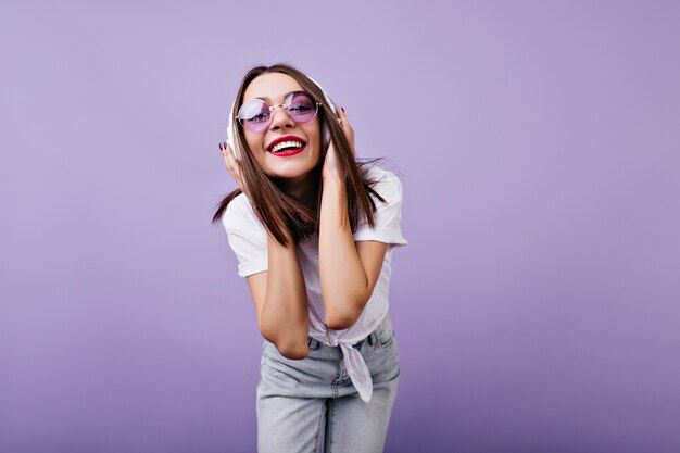 Blij meisje in zonnebril haar koptelefoon met glimlach aan te raken. Binnenfoto van schitterend vrouwelijk model in geïsoleerde jeans.