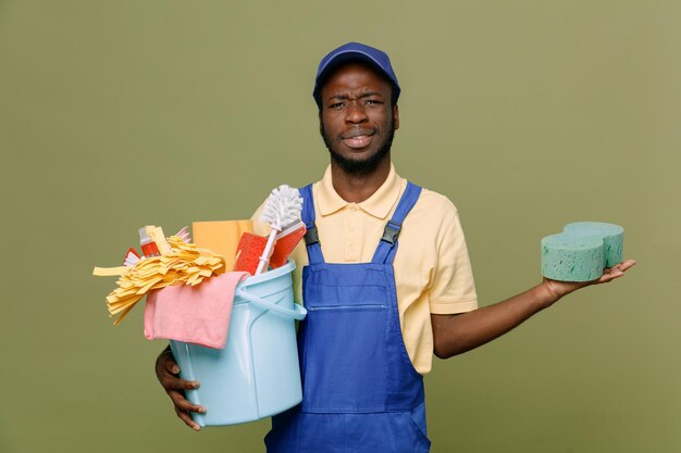 Blij met emmer schoonmaakgereedschap met spons jonge Afro-Amerikaanse schonere man in uniform met handschoenen geïsoleerd op groene achtergrond