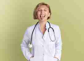 Gratis foto blij met gesloten ogen jonge vrouwelijke arts die medische mantel draagt met een stethoscoop die tong toont geïsoleerd op olijfgroene muur