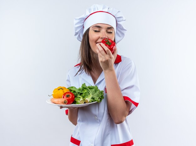 blij met gesloten ogen jonge vrouwelijke kok in uniform van de chef-kok met groenten op bord snuivende peper geïsoleerd op een witte muur