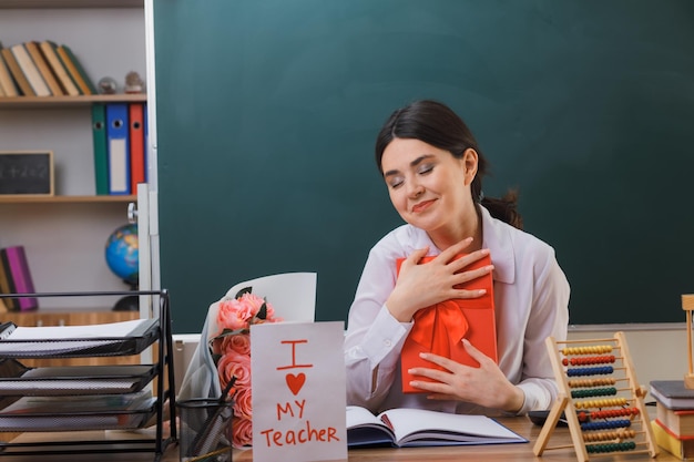 Blij met gesloten ogen jonge vrouwelijke leraar met cadeau zittend aan bureau met schoolhulpmiddelen in de klas