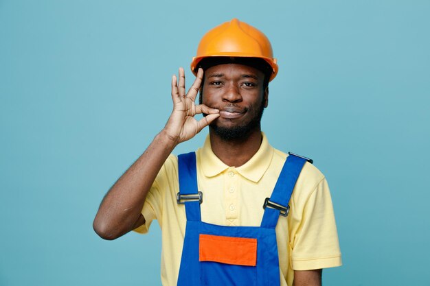 Blij met heerlijk gebaar jonge Afro-Amerikaanse bouwer in uniform geïsoleerd op blauwe achtergrond