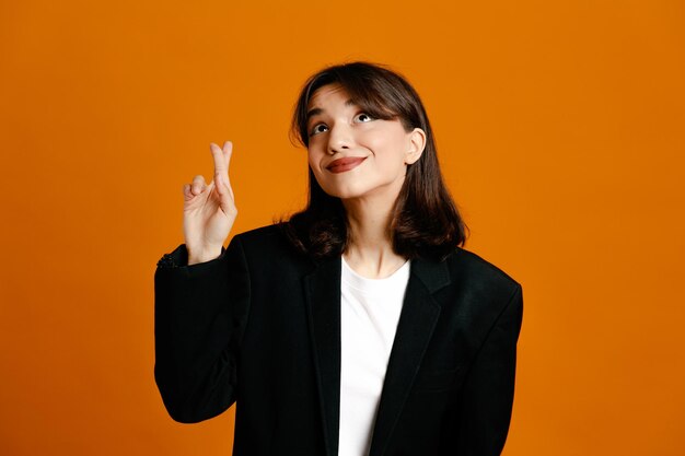 Blij met het kruisen van vingers jonge mooie vrouw met zwarte jas geïsoleerd op een oranje achtergrond