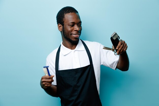 Blij met kapper tools jonge Afro-Amerikaanse kapper in uniform geïsoleerd op blauwe achtergrond