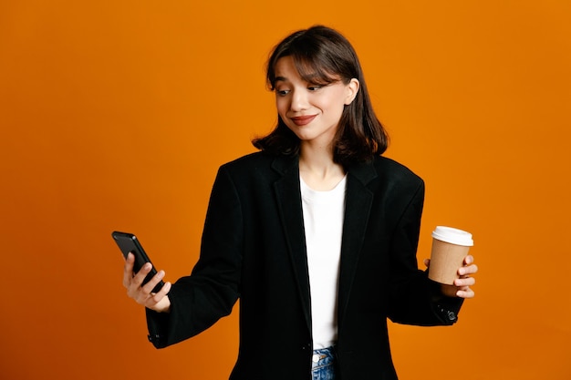 Blij met koffiekopje en telefoon jonge mooie vrouw met zwarte jas geïsoleerd op oranje achtergrond