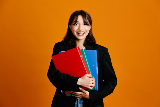 Blij met mappen jonge mooie vrouw met zwarte jas geïsoleerd op een oranje achtergrond