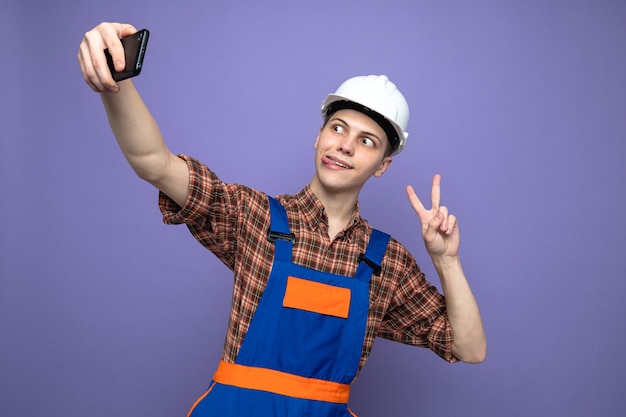 Blij met tong en vredesgebaar, jonge mannelijke bouwer die uniform draagt, neemt een selfie