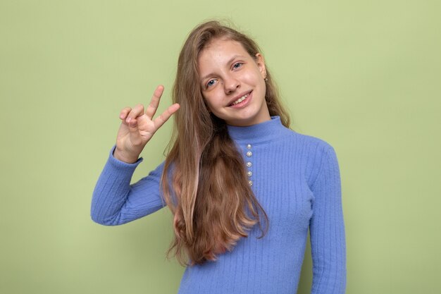 Blij met vredesgebaar mooi klein meisje met blauwe trui geïsoleerd op olijfgroene muur