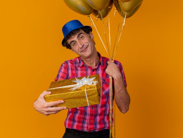 Blij middelbare leeftijd blanke partij man met feestmuts houden ballonnen kijken camera strekken cadeaupakket naar camera geïsoleerd op een oranje achtergrond met kopie ruimte