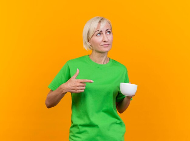 Blij middelbare leeftijd blonde Slavische vrouw met kopje thee kijken en wijzend naar kant geïsoleerd op gele muur met kopie ruimte