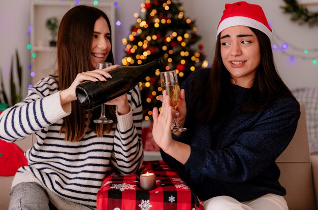 Blij mooi jong meisje met kerstmuts houdt glas champagne en gebaren stopbord naar haar vriend fles champagne zittend op een stoel te houden en thuis te genieten van de kersttijd