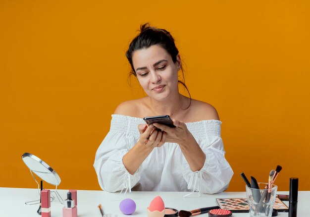 Blij mooi meisje zit aan tafel met make-up tools houden en kijken naar telefoon geïsoleerd op oranje muur