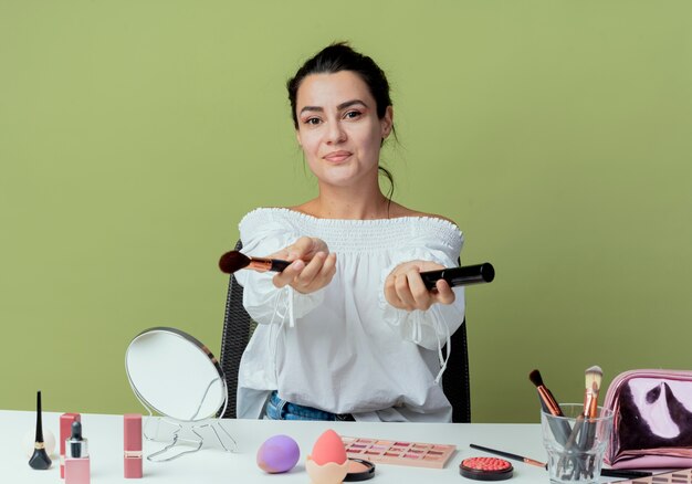 Blij mooi meisje zit aan tafel met make-up tools houdt make-up borstel en mascara op zoek geïsoleerd op groene muur