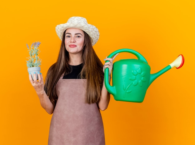 Blij mooi tuinman meisje dragen uniform en tuinieren hoed bedrijf gieter met bloem in bloempot geïsoleerd op een oranje achtergrond