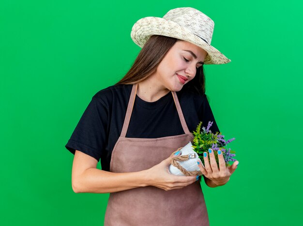 Blij mooi tuinman meisje in uniform dragen tuinieren hoed bedrijf en kijken naar bloem in bloempot geïsoleerd op groene achtergrond