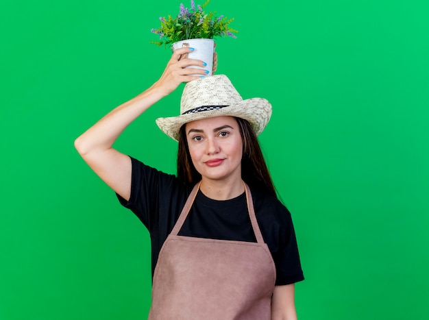 Gratis foto blij mooi tuinman meisje in uniform dragen tuinieren hoed met bloem in bloempot op hoofd geïsoleerd op groene achtergrond