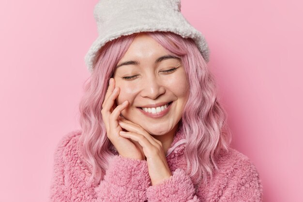 Blij mooie Aziatische vrouw raakt gezicht zachtjes houdt ogen gesloten glimlacht tandjes dagdromen over iets draagt bontjas en hoed geïsoleerd over roze achtergrond Oprechte emoties en gevoelens