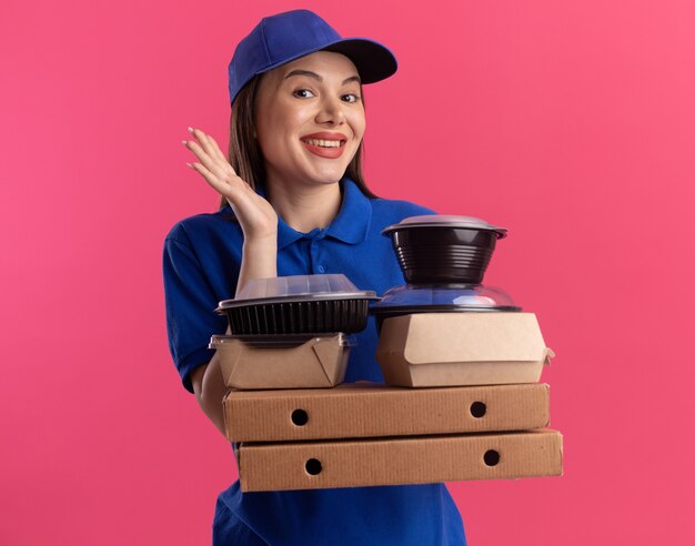 Blij mooie bezorger in uniform staat met opgeheven hand en houdt voedselpakket en containers op pizzadozen
