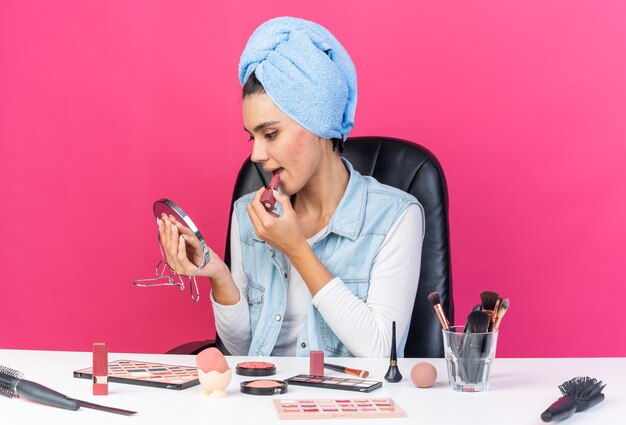 Blij mooie blanke vrouw met gewikkeld haar in een handdoek zittend aan tafel met make-up tools houden en kijken naar spiegel lippenstift aan te brengen