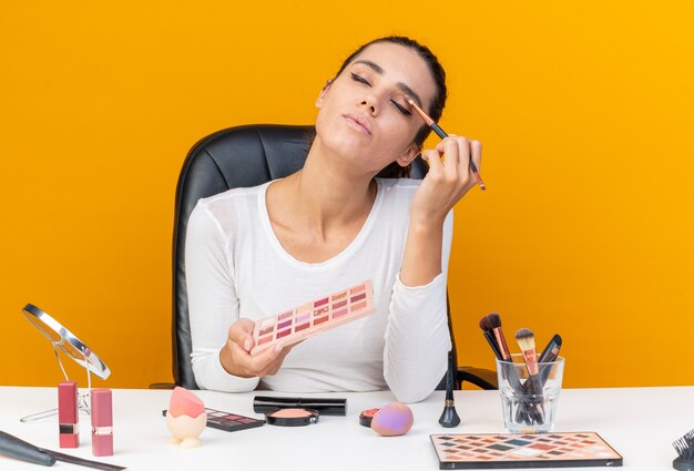 Blij mooie blanke vrouw zittend aan tafel met make-up tools houden oogschaduw palet en het toepassen van oogschaduw geïsoleerd op oranje muur met kopie ruimte