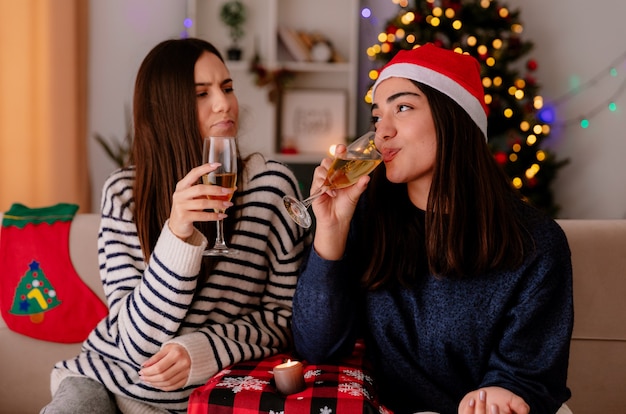 blij mooie jonge meisjes drinken van glazen champagne zittend op fauteuils en genieten van kersttijd thuis