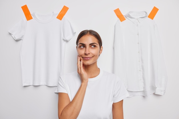 Blij nadenkende Europese vrouw heeft een dromerige uitdrukking, denkt aan iets houdt de hand op het gezicht poses tegen een witte achtergrond gepleisterde kleding achter overweegt wat te dragen. Laat me denken