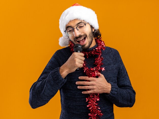 Blij neerkijkt jonge knappe kerel met kerstmuts met slinger op nek spreekt op microfoon hand op zichzelf geïsoleerd op een oranje achtergrond