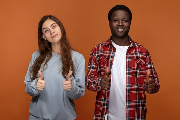 Blij om samen te zijn. Foto van positieve vrolijke jonge Afro-Amerikaanse jongen en zijn schattige langharige Kaukasische vriendin genieten van tijd samen, vreugdevol glimlachen en duimen opdagen gebaar