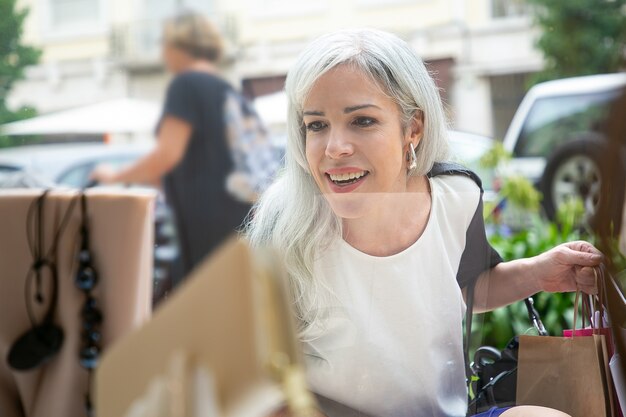 Blij opgewonden shopaholic staren naar accessoires in de etalage, boodschappentassen vasthouden, staande in de winkel buiten. Vooraanzicht door glas. Window shopping concept