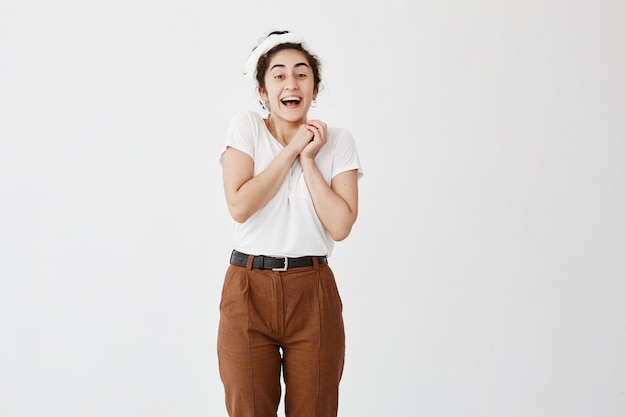 Blij opgewonden vrouw met donker haar in knot, draagt een wit t-shirt, ziet er met geluk uit, lacht breed met geopende mond, houdt de handen bij elkaar op de borst. geluk en schoonheid concept