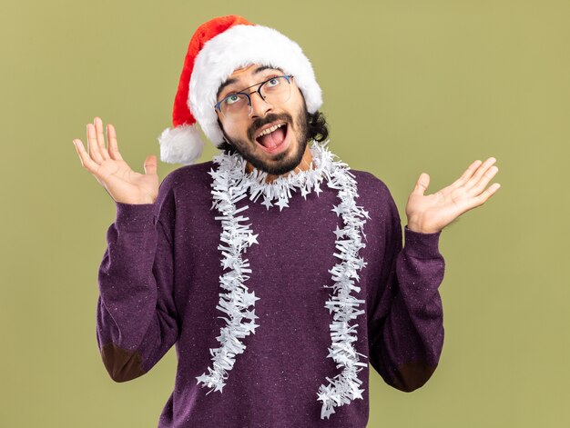 Blij opzoeken van jonge knappe man met kerstmuts met slinger op nek spreidende handen geïsoleerd op olijfgroene achtergrond