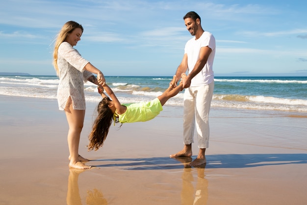 Blij paar en meisje die pret ionenstrand hebben, ouders die meisjesarmen en benen houden en haar wiegen