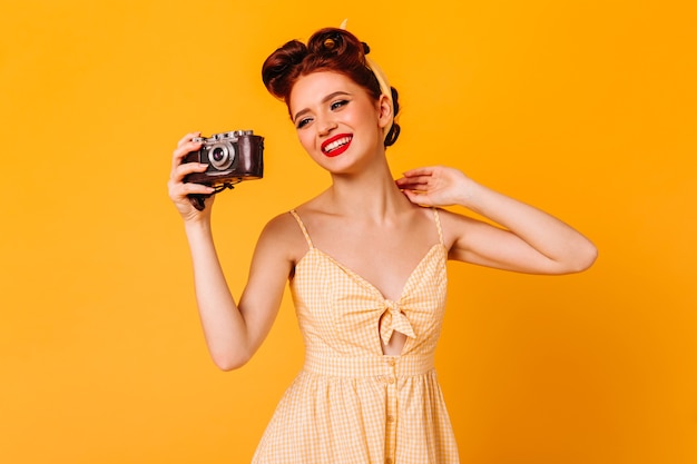 Gratis foto blij pinupmeisje die in kleding foto's nemen. studio shot van elegante lachende vrouw met camera geïsoleerd op gele ruimte.