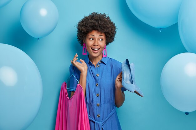 Blij positief Afro-Amerikaanse dame kiest outft voor date, houdt blauwe schoenen met hoge hakken en roze jurk aan hangers vast, bereidt zich voor op feest en feest, poseert over blauwe muur met opgeblazen ballonnen