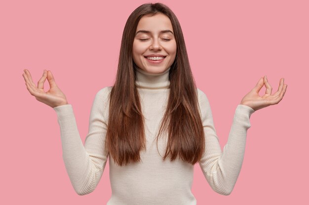 Blij positieve jonge Europese vrouw maakt mudra gebaar, houdt de ogen dicht, ontspant na het werk, gekleed in vrijetijdskleding, geïsoleerd op roze achtergrond. Mensen, welzijn