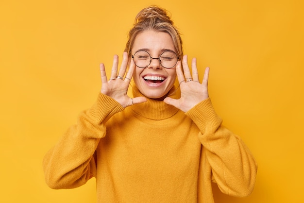 Blij positieve jonge vrouw houdt handpalmen omhoog in de buurt van gezicht roept vrolijk sluit ogen draagt casual jumper glimlacht breed toont witte tanden geïsoleerd over gele achtergrond Menselijke emoties concept