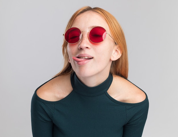 Blij roodharig gembermeisje met sproeten in zonnebril steekt tong uit geïsoleerd op een witte muur met kopieerruimte