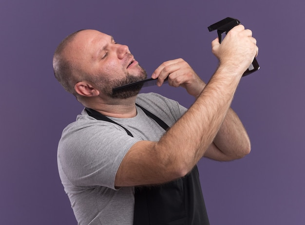 Blij Slavische mannelijke kapper van middelbare leeftijd in uniform baard kammen en water geven met spuitfles geïsoleerd op paarse muur