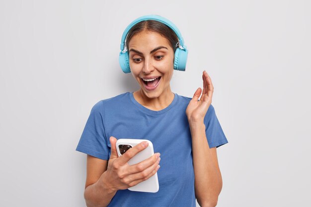 Blij verrast donkerharige jonge vrouwelijke model gericht op smartphone downloads liedjes naar afspeellijst draagt stereo koptelefoon op oren gekleed in casual blauw t-shirt geïsoleerd op witte achtergrond