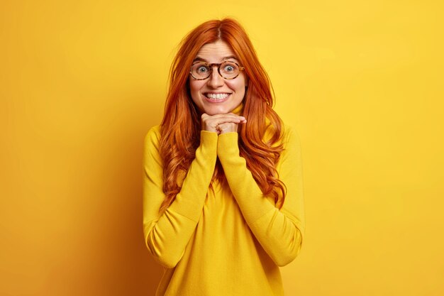 Blij verrast roodharige jonge vrouw houdt handen onder kin toont witte tanden voelt blij luistert met grote belangstelling gesprekspartner gekleed in casual trui.