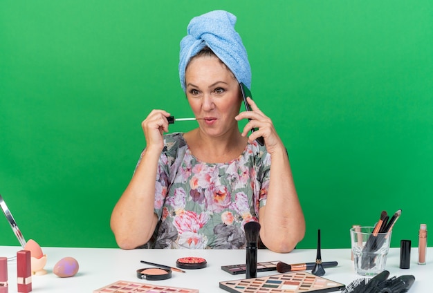 Blij volwassen blanke vrouw met gewikkeld haar in een handdoek zittend aan tafel met make-up tools praten over de telefoon lipgloss toe te passen