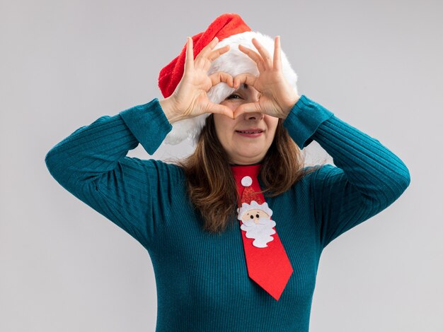 Blij volwassen blanke vrouw met kerstmuts en santa stropdas gebaren en door hart teken geïsoleerd op een witte muur met kopie ruimte